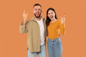 A joyful couple enthusiastically makes rock-on hand gestures while smiling widely. The background is a vibrant orange color, highlighting their cheerful expressions and casual attire. photo