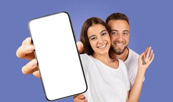 A young couple stands in front of a solid blue background, smiling brightly at the camera. The woman is holding a smartphone with a blank screen photo