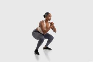 A focused African American woman athlete is engaged in a strength-building exercise routine, demonstrating perfect form while doing squats on white background photo