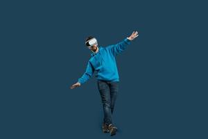A young man engages in a balance exercise while wearing a virtual reality headset. He stands on one leg, arms outstretched for stability, in a modern indoor setting with a solid color backdrop photo