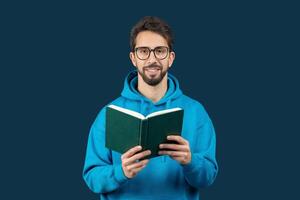 A man wearing a blue hoodie is engrossed in reading a book. He sits indoors, focused on the pages, with a calm demeanor as he flips through the book. photo