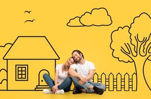 A joyful couple sit closely together, leaning on each other with a contented embrace against a playful backdrop that includes a sketched house, tree, clouds, and birds on a vibrant yellow background photo