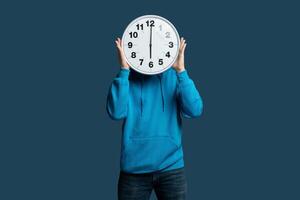 A young man stands against a blue wall, holding a large wall clock in front of their face. He is dressed in a blue hoodie and jeans, creating a playful and humorous visual representation of time photo