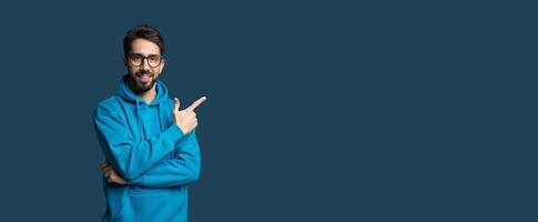 A young man wearing a blue hoodie stands confidently with his arms crossed and a smile on his face. He points at copy space, engaging viewers attentively. The background is a simple, solid color photo