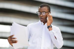 A young black businessman stands outside a contemporary building, attentively holding several documents in one hand. He is using his smartphone to have an important conversation photo