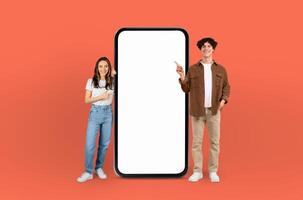 A man and woman are standing in front of a blank screen, both pointing towards it, against a vibrant orange background. They seem to be discussing or presenting something on the screen. photo