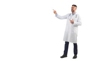 A man doctor is standing against a plain white background while pointing to the side and smiling. He is wearing a white lab coat with a stethoscope around his neck, copy space photo