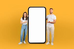 A man and woman are standing beside a blank phone screen, posing for a photo. They both appear to be smiling and looking at the screen, possibly preparing to take a selfie or share something photo