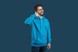 A cheerful guy wearing a bright blue hoodie stands confidently, flashing a warm smile. He makes a playful calling gesture with his hand near his ear photo