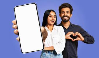 A young Indian couple stands in front of a blue background. The woman holds a smartphone with a blank screen in her left hand and points to it with her right. The man is making a heart shape gesture photo
