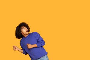 A young guy with curly hair displays a surprised expression, appearing to run away from something off-screen. The vibrant yellow background adds a sense of excitement to the moment, copy space photo