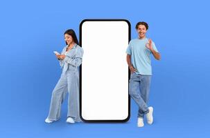 A man and a woman are standing side by side, looking at a blank screen of a phone in a neutral environment. They seem to be discussing or viewing something on the screen photo