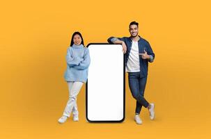 Multiracial man and woman standing in front of blank phone screen, striking pose and looking directly at camera. Phone screen is void of any content, emphasizing duos interaction with technology. photo