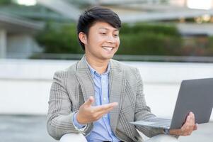 Asian young man is sitting outside on a bright day and smiling while using his laptop computer. He is wearing a plaid blazer, having a conversation, possibly over a call. photo