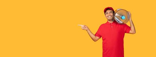 Delivery man wearing a red uniform is standing over yellow studio background while holding a large bottle of water in his hand, panorama with copy space photo