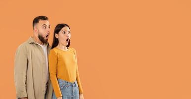 A young couple stands close together, displaying shocked expressions with wide-open mouths, against a solid orange background, panorama with copy space photo