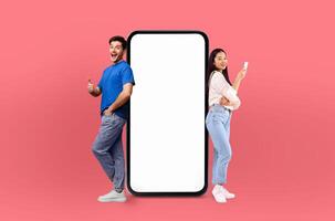 Multiethnic lovers stand on either side of a large smartphone, both expressing excitement and approval with thumbs up gestures photo