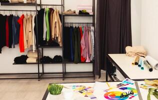 A room featuring a wooden table with a stack of fabric and a metal rack filled with various clothes such as jackets, shirts, and dresses. The setting appears tidy and functional, designer workspace photo