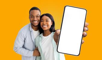A smiling black couple holding a smartphone with a white blank screen in front of them against a bright yellow background. The woman is pointing at the screen, and both are looking at the camera. photo