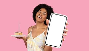 African American young woman with curly hair is holding a piece of birthday cake in one hand and a smartphone with a blank screen in the other hand. The background is a solid pink color. photo