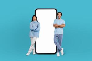 On a deep blue background, a multiethnic couple, both smiling, stands adjacent to an enormous smartphone with a blank display, ready for showcasing apps or designs. photo
