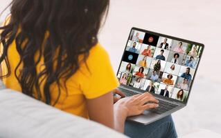 Black woman with long, black hair sits on a couch while participating in an online meeting, using a laptop with a screen showing a grid of multiple feeds of other participants in the meeting. photo