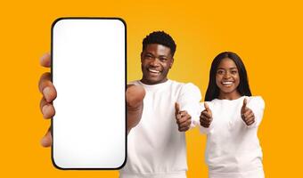 A young man and woman, both African American and smiling, stand side-by-side in front of a bright yellow background. The man holds a smartphone with a blank white screen photo