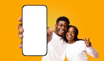 A young black couple smiles brightly as they hold a smartphone with a blank white screen in front of them. The woman makes a peace sign with her hand, while the man points at the screen. photo