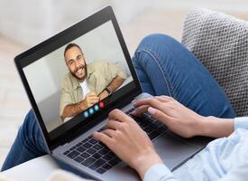 A woman is sitting on a couch and chatting with a man on a laptop computer. She is typing on the keyboard and looking at the screen. The man is smiling and appears to be talking. photo