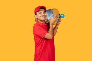A cheerful young delivery man in a red uniform and cap holds a large water container on his shoulder. The bright yellow background adds a vibrant feel to the scene photo