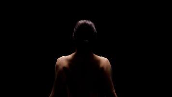 A woman sits with her back to the camera, illuminated by a single light source. She is facing away from the viewer and the background is completely black. photo