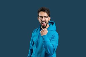 un vibrante joven hombre con lentes es capturado A mitad de la ovación, vistiendo un brillante azul sudadera. él aprieta su puño y abre su boca amplio en un gesto de emoción o apoyo foto