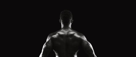 A muscular man stands with his back to the camera in a dark studio, lit by a single light source. photo