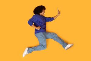 A man is mid-air, captured while jumping energetically with one hand holding a cell phone. His body is in motion, creating a dynamic and active scene. photo