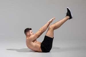 A shirtless man demonstrates the hollow body hold exercise while wearing black shorts and sneakers. The studio has a plain gray backdrop, emphasizing the form and position of the exercise. photo