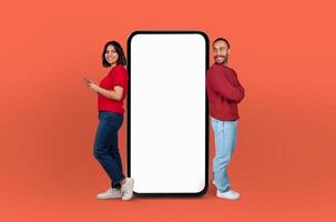 Multiethnic couple stand on either side of a giant, blank smartphone screen, both smiling and engaging with the empty display photo