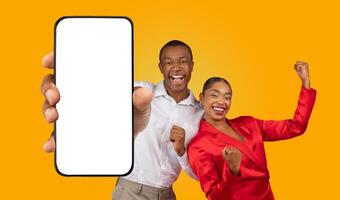 African American happy couple standing against a yellow background. The man is holding a smartphone with a blank screen in front of him and pointing at it with his right hand. photo