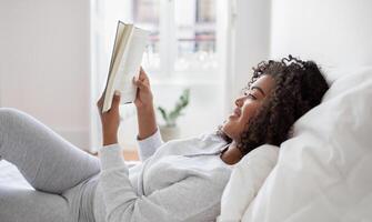A Hispanic woman is lying down in bed with a book in hand, engrossed in reading. The room is dimly lit, and she appears relaxed and focused on the pages of the book, side view photo