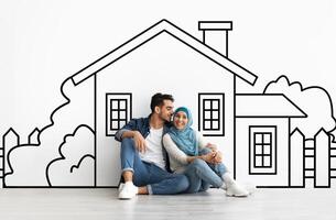 Loving millennial muslim couple planning their dream home against a hand-drawn house sketch on white wall background. Mortgage concept photo