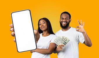 A happy black couple poses against a vibrant yellow backdrop. The woman showing smartphone in her hand, displaying a blank screen, while the man holds a fan of cash and makes the okay gesture photo