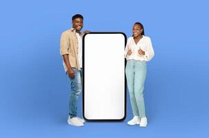 A young black man and woman are standing next to a blank screen in front of a blue background, dressed casually and appear to be engaged in conversation or viewing something on the screen. photo