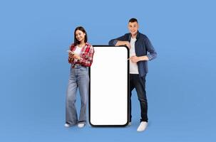Multiethnic man and woman are standing next to a blank screen. The man is wearing a blue shirt and the woman a black dress. Both are looking at the screen with serious expressions. photo