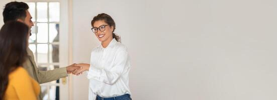 A businesswoman wearing glasses and a white shirt is shaking hands with a client in a modern office setting. The clients, who is standing next to another person, is smiling and seems happy, copy space photo