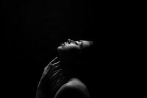 A black and white photo of a woman with her eyes closed, reaching her hands upwards. Her face is lit by a single light source from above, creating a dramatic contrast against the dark background.