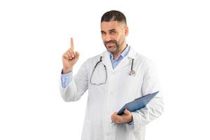 A man doctor dressed in a white lab coat stands confidently with one hand holding a clipboard and the other hand pointing upward. The background is a clean, white studio setting photo