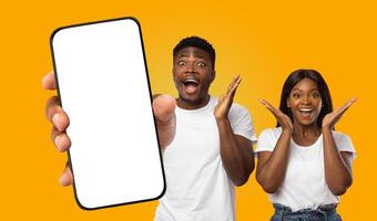 Black couple standing in front of a yellow background, looking at a blank smartphone screen being held out by a hand. Both have their hands raised in excitement with wide smiles on their faces. photo