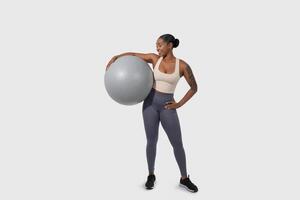 African American woman is holding an exercise ball in front of a plain white background. She stands confidently, gripping the ball with both hands, with a focused expression. photo