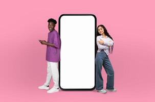 Illuminated by the cool pink backdrop, the multiethnic young man and woman share a smile, enhancing the visual appeal of the blank smartphone screen beside them. photo