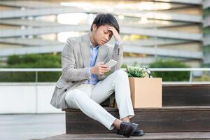 Asian young man in a blazer and white pants sits on a set of stairs, holding his phone and looking down at it with a worried expression. He has a cardboard box beside him. photo