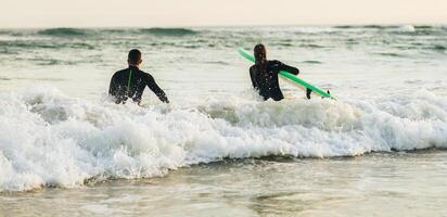 This image shows two surfers entering the ocean at sunset. They are wearing wetsuits and carrying surfboards. The waves are breaking in the background and the sky is beautiful photo